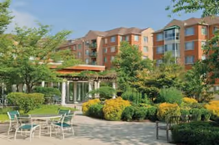 Sunny courtyard with patio tables, landscaped shrubs and a gazebo in front of a multi-story senior living building.