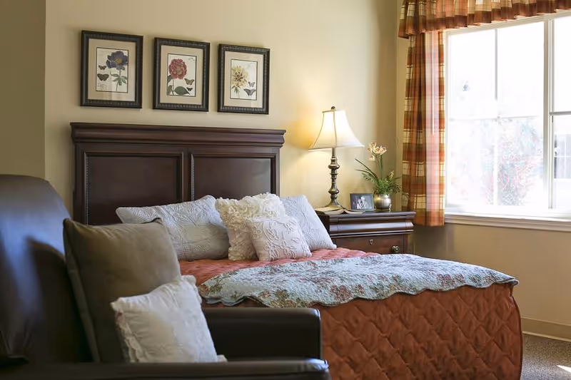 A cozy bedroom with a large bed featuring multiple pillows and a quilted bedspread. There is a dark wooden headboard and a matching nightstand with a lamp, a small plant, and a framed photo. Three framed floral prints hang on the wall above the bed. A window with plaid curtains allows natural light to brighten the room. In the foreground, there is a dark leather armchair with pillows.