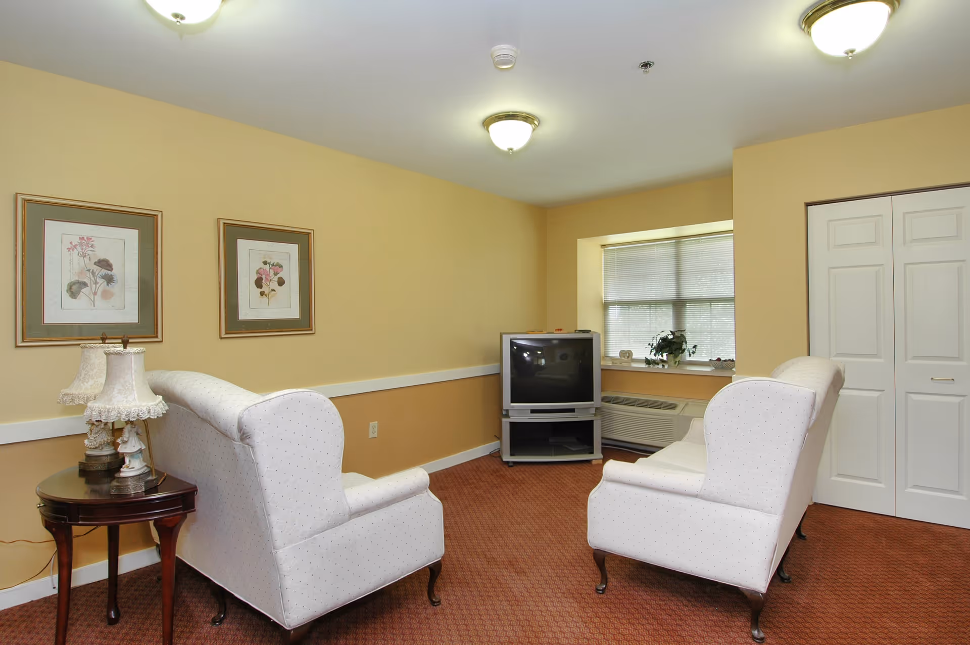 A small living room with two white upholstered armchairs facing an older model television on a stand. The walls are painted beige with a white chair rail, and there are two framed floral prints hanging on the wall. A small wooden side table with a decorative lamp is next to one of the chairs. A window with blinds and a potted plant is visible behind the TV, and there is a white closet door on the right side of the room. The carpet is a reddish-brown color.