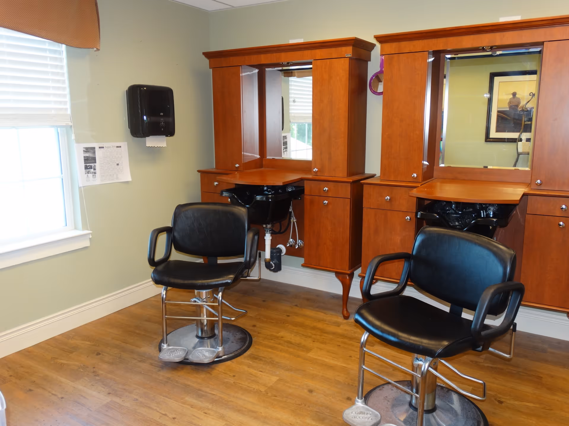 Interior view of a salon area with two black salon chairs in front of wooden cabinetry with mirrors and built-in sinks. A window with blinds and a paper towel dispenser are visible on the left wall.