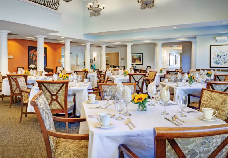 Formal dining room with multiple tables set with white linens, place settings, glassware, and sunflower centerpieces.
