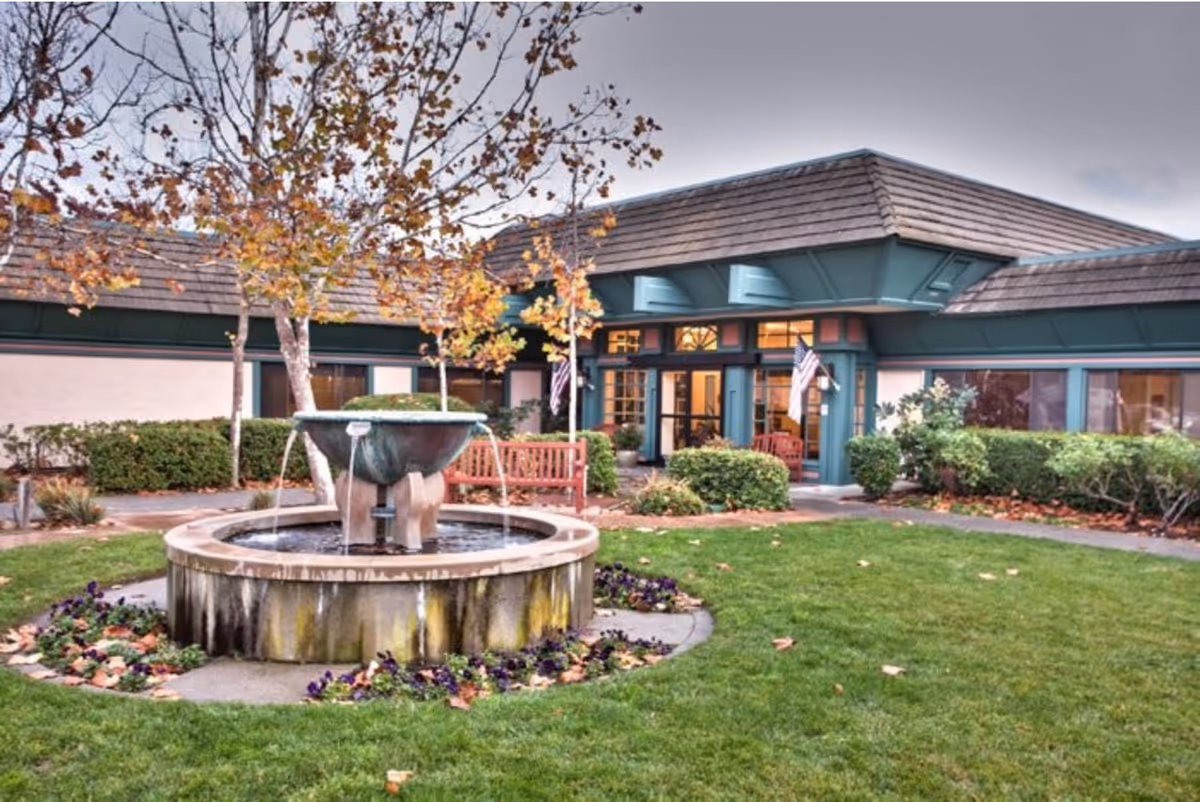 Outdoor courtyard area of Walnut Creek Skilled Nursing & Rehabilitation Center featuring a circular water fountain surrounded by flowers and green grass. There are benches and bushes around the courtyard, with the building's entrance visible in the background under a cloudy sky.