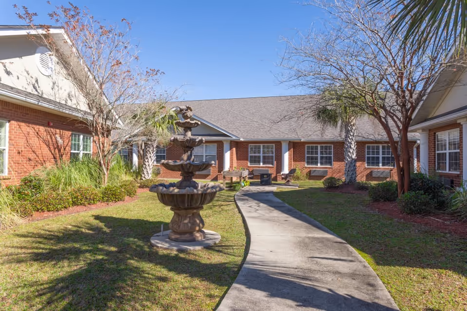 Sunlit courtyard with a multi-tiered fountain, curved concrete pathway, benches, and surrounding brick senior living buildings.