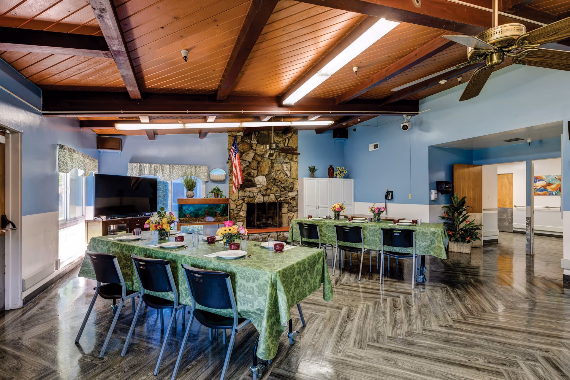 A dining room with two tables covered in green patterned tablecloths, set with plates, cups, and flower centerpieces. The room has a wooden ceiling with exposed beams, a ceiling fan, a stone fireplace, a television, and an aquarium. The walls are painted light blue with white trim, and there are windows with floral valances letting in natural light.