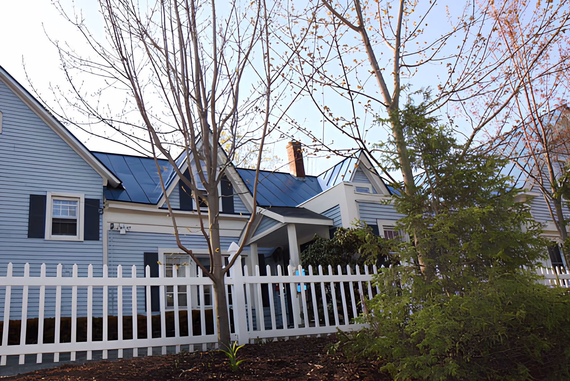 Front exterior of a light blue house with a white picket fence, trees, and landscaping.