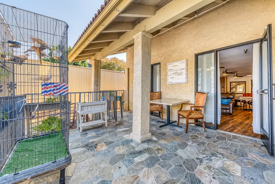 Outdoor patio area with stone tile flooring, two upholstered chairs and a table near the wall, a large birdcage on the left side, and an open sliding glass door leading to an interior living room with wooden floors and furniture.
