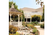Outdoor garden area with a white gazebo, a small rock-lined water feature, shrubs, and a paved walkway leading through the landscaped grounds. A building with beige siding and a brown roof is visible in the background.