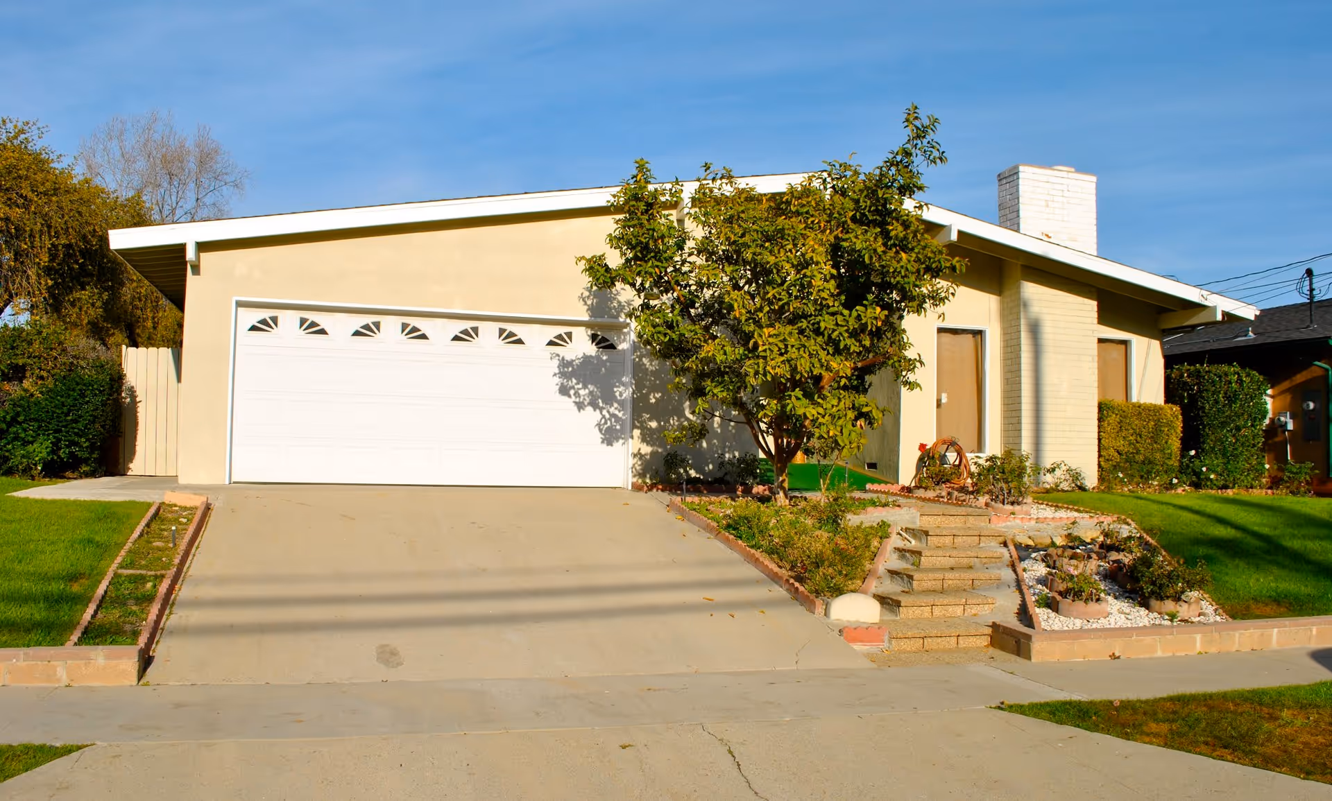 Exterior view of a single-story residential building with a white garage door, a driveway, a small garden with a tree, and steps leading up to the front door under a clear blue sky.