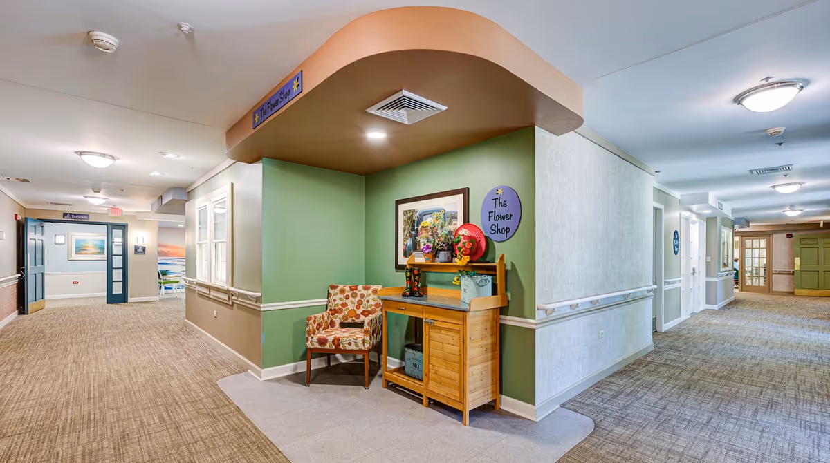 Interior hallway of Arden Courts - ProMedica Memory Care Community with beige and green walls, carpeted floors, ceiling lights, and a small wooden display labeled 'The Flower Shop' featuring flowers and decorative items next to a floral-patterned armchair.