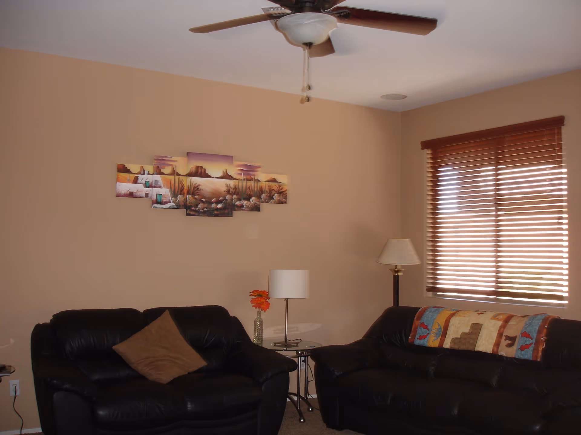 A living room with two black leather sofas, one with a brown pillow and the other with a colorful quilt draped over the back. There is a small glass side table with a lamp and a vase with orange flowers between the sofas. A floor lamp stands next to the sofa on the right. The walls are beige, and there is a multi-panel desert landscape painting hanging on the wall. A window with wooden blinds is on the right side, and a ceiling fan with a light fixture is mounted on the white ceiling.