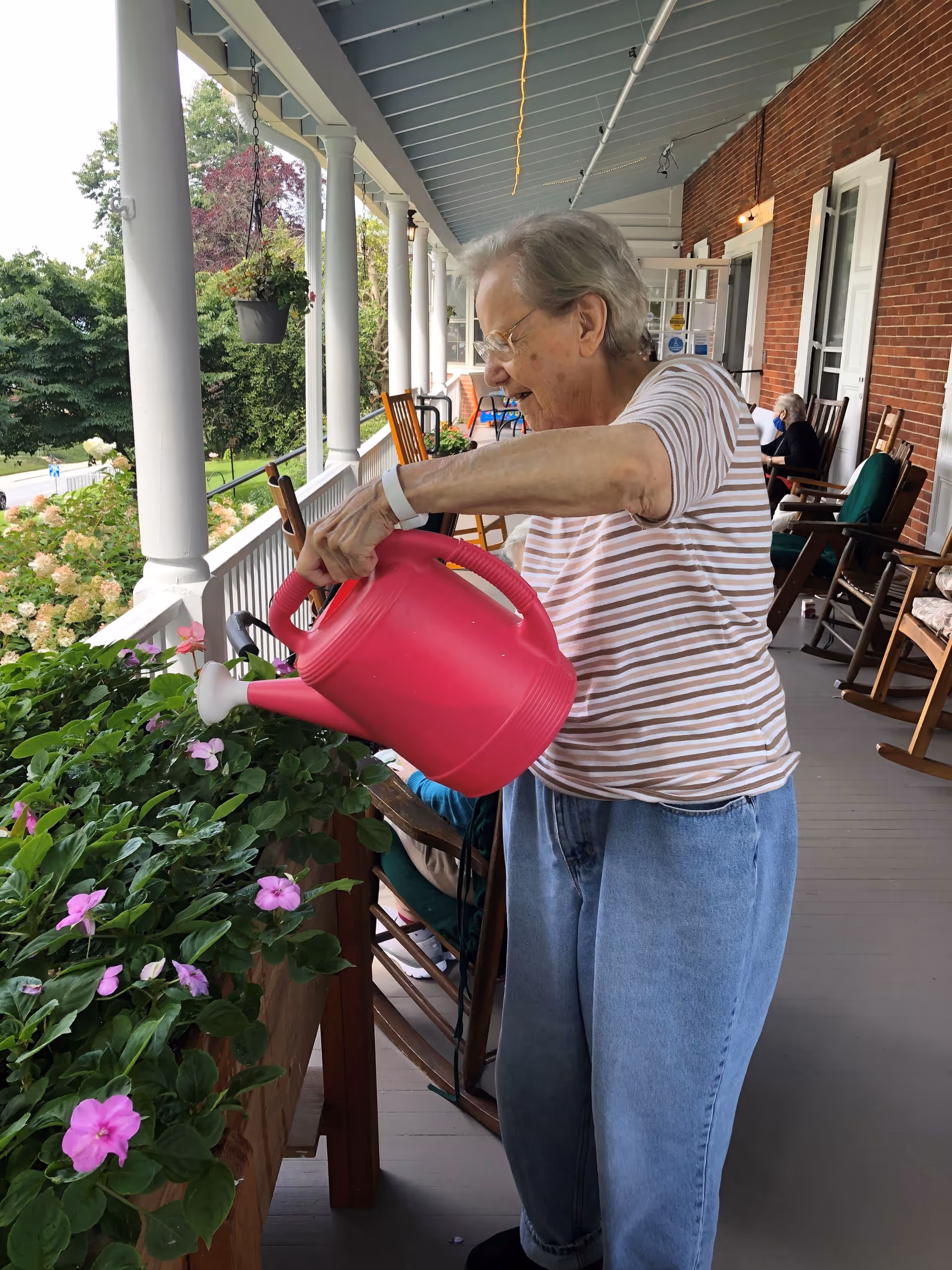 An elderly woman wearing glasses, a striped shirt, and blue jeans is watering pink flowers on a porch with a bright pink watering can. The porch has white columns, wooden rocking chairs, and hanging plants, with greenery and trees visible in the background.