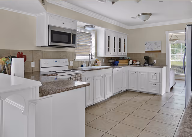 A bright and clean kitchen with white cabinets, a white stove, a microwave above the stove, a sink under a window, and various kitchen items on the countertops. The floor is tiled, and there is a doorway leading to another room with a window and a chandelier.