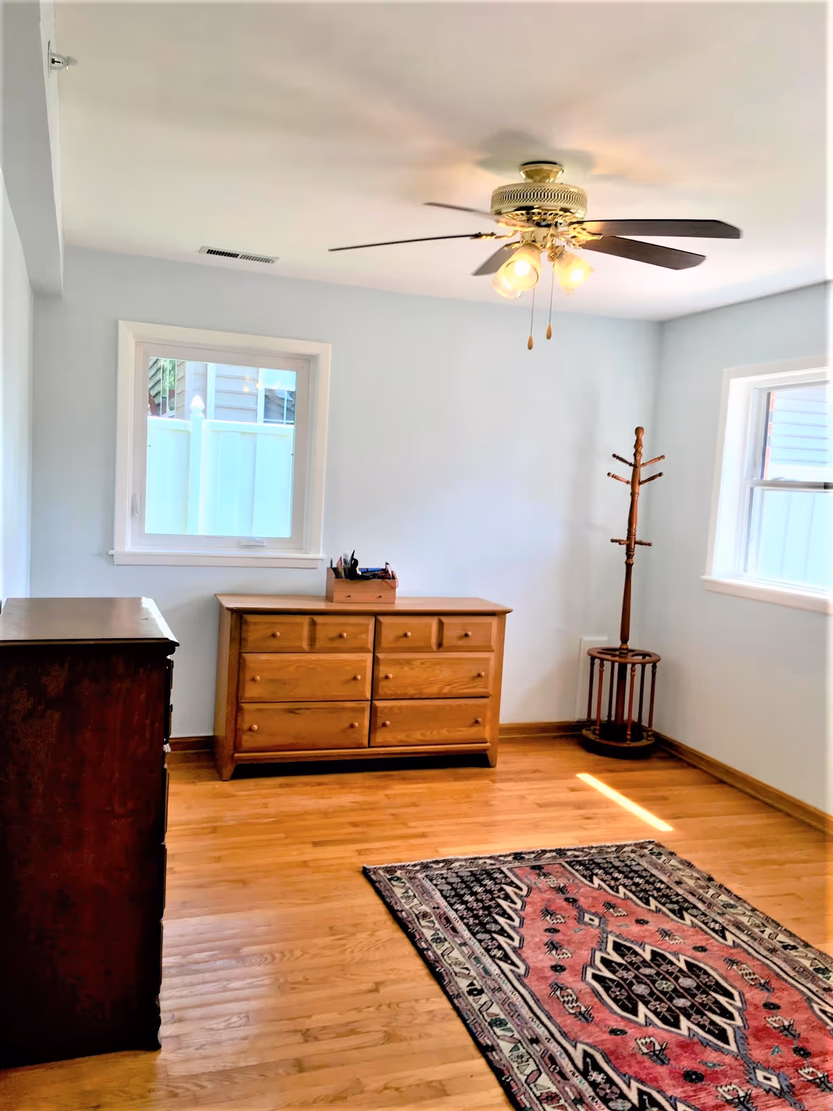 A bright room with light blue walls, two windows, a ceiling fan with lights, a wooden dresser, a wooden chest of drawers, a wooden coat rack, and a patterned area rug on a hardwood floor.