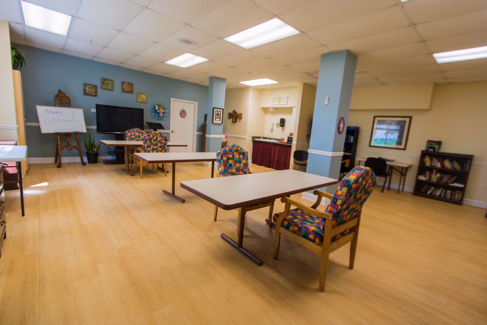 A spacious room with light wooden flooring and a drop ceiling with fluorescent lights. The room contains several tables with colorful patterned chairs around them. There is a large flat-screen TV mounted on a blue accent wall, a whiteboard on an easel with writing on it, and some framed pictures and decorations on the walls. In the background, there is a small kitchenette area with a counter and a sink. A bookshelf and a desk with a chair are visible on the right side of the room.