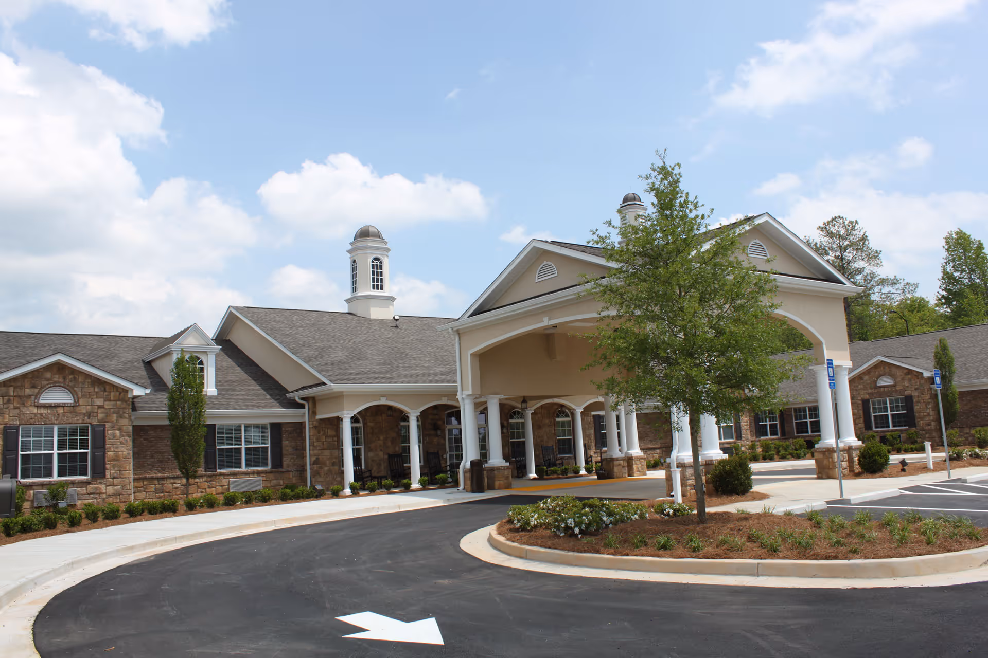 Exterior view of Benton House of Newnan, showing a large building with stone and beige siding, a covered entrance with white columns, a circular driveway with a white arrow painted on the asphalt, landscaped areas with bushes and a tree, and a partly cloudy sky.