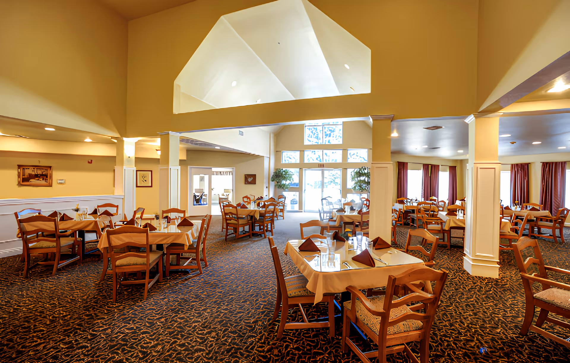 Spacious dining room with multiple wooden tables and chairs arranged neatly. Tables are covered with beige tablecloths and set with folded brown napkins, glasses, and silverware. The room features a high ceiling with a large triangular window letting in natural light, beige walls, patterned carpet, and several framed pictures on the walls. Large windows with red curtains are visible on the right side.