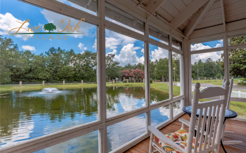 View from a screened porch with a white rocking chair and small round table, overlooking a pond with a fountain and surrounded by trees under a blue sky with clouds.