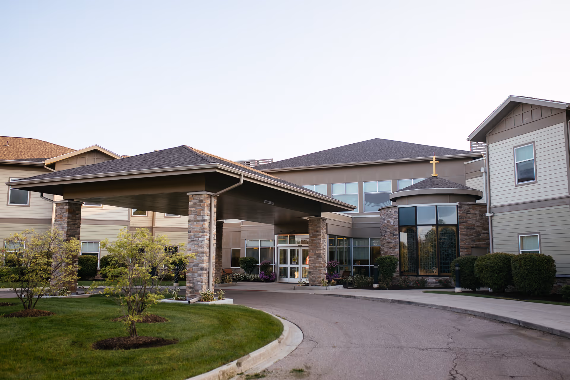 Exterior view of The Neighborhoods of White Lake senior living facility entrance with a covered driveway supported by stone pillars, manicured green lawn, small trees, and bushes under a clear sky.