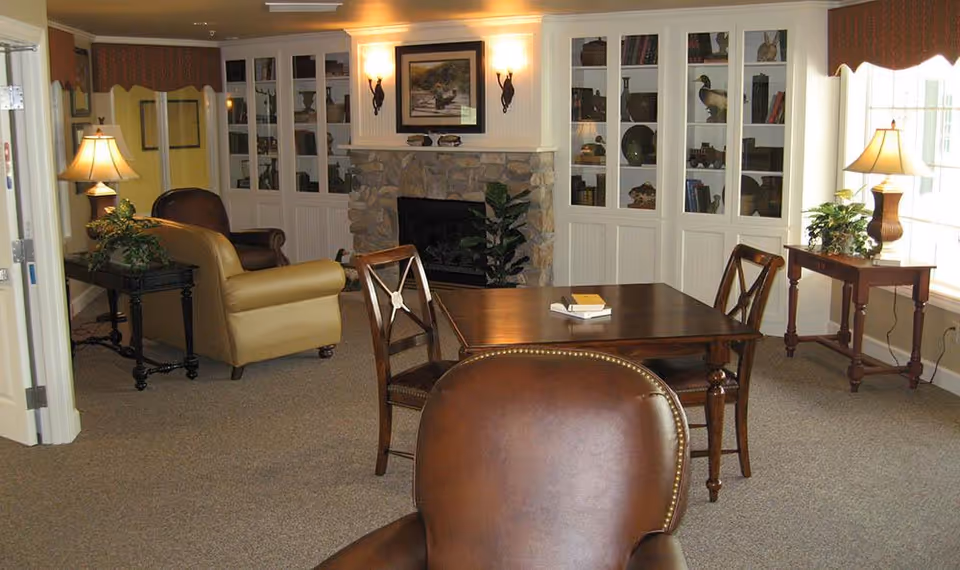 Cozy common room with leather armchairs, a wooden table and chairs, built-in bookcases flanking a stone fireplace, lamps, and windows.
