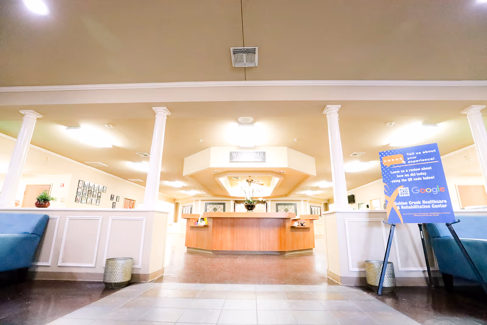 Reception area of Golden Creek Healthcare and Rehabilitation with a wooden front desk in the center, white columns, beige walls, blue chairs on either side, and a blue sign on an easel encouraging visitors to leave a Google review.