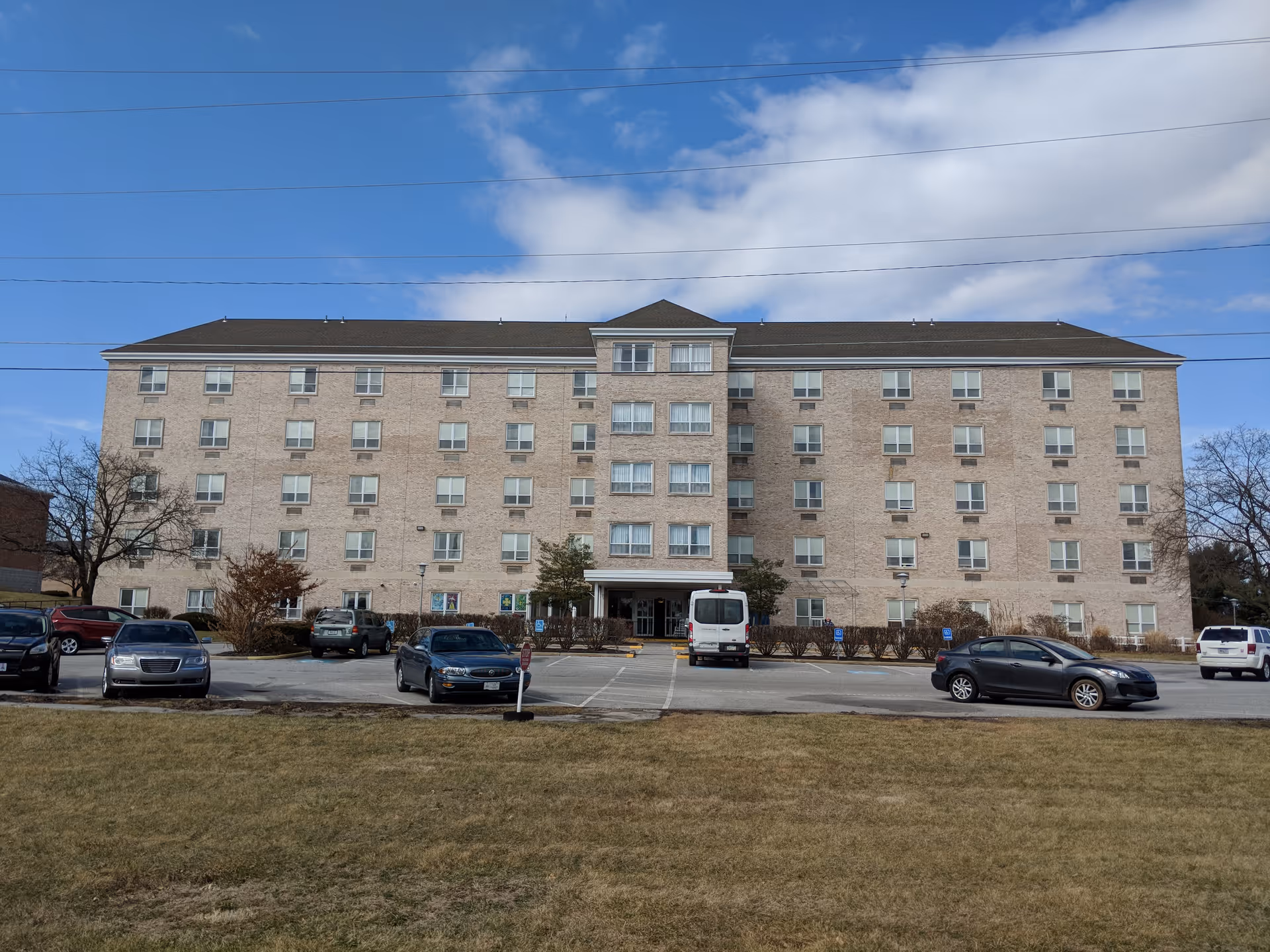 Front exterior of a five-story brick nursing and rehabilitation building with a parking lot and several cars.