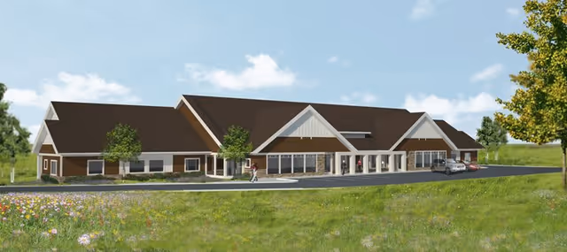 Front-view rendering of a single-story senior living building with a pitched roof, covered entrance, driveway with cars, and surrounding lawn.