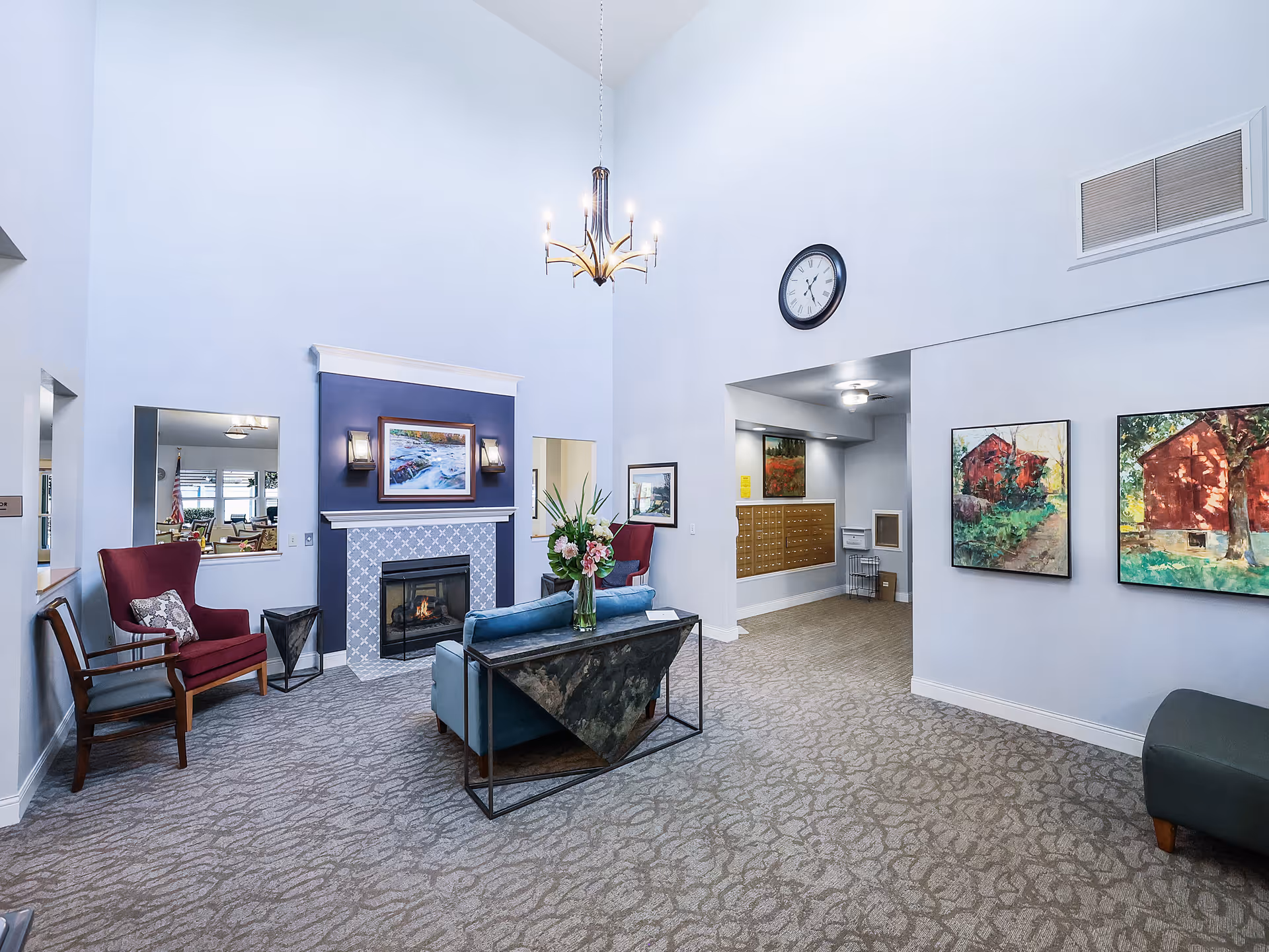 A spacious senior living facility common area with high ceilings, a chandelier, a fireplace with a painting above it, two red armchairs, a blue sofa with a floral arrangement on a table behind it, carpeted floor, wall clock, and colorful paintings on the walls.