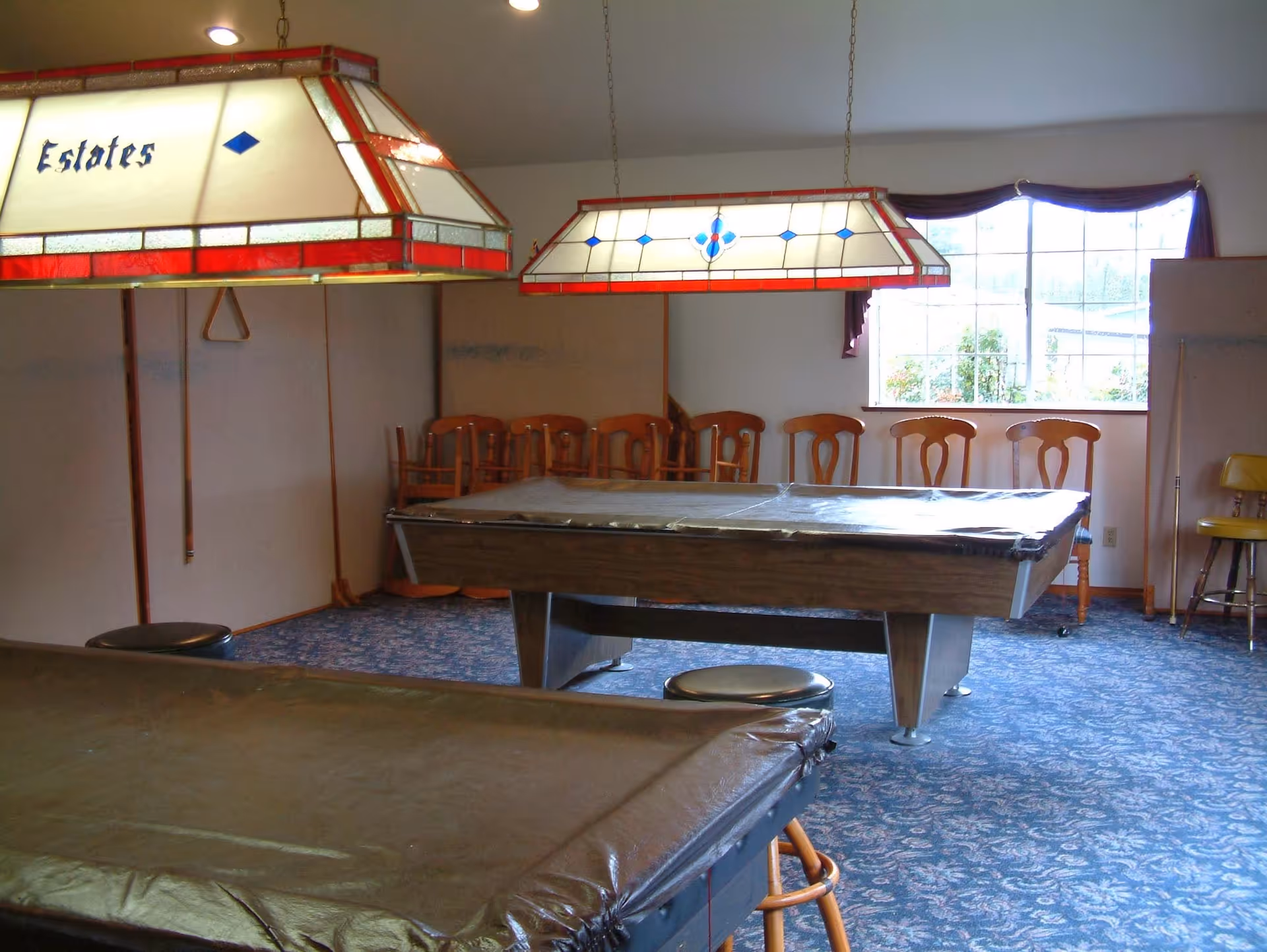 A recreational room with two covered pool tables, wooden chairs lined up against the wall, a window with a valance, and two hanging stained glass light fixtures above the pool tables.