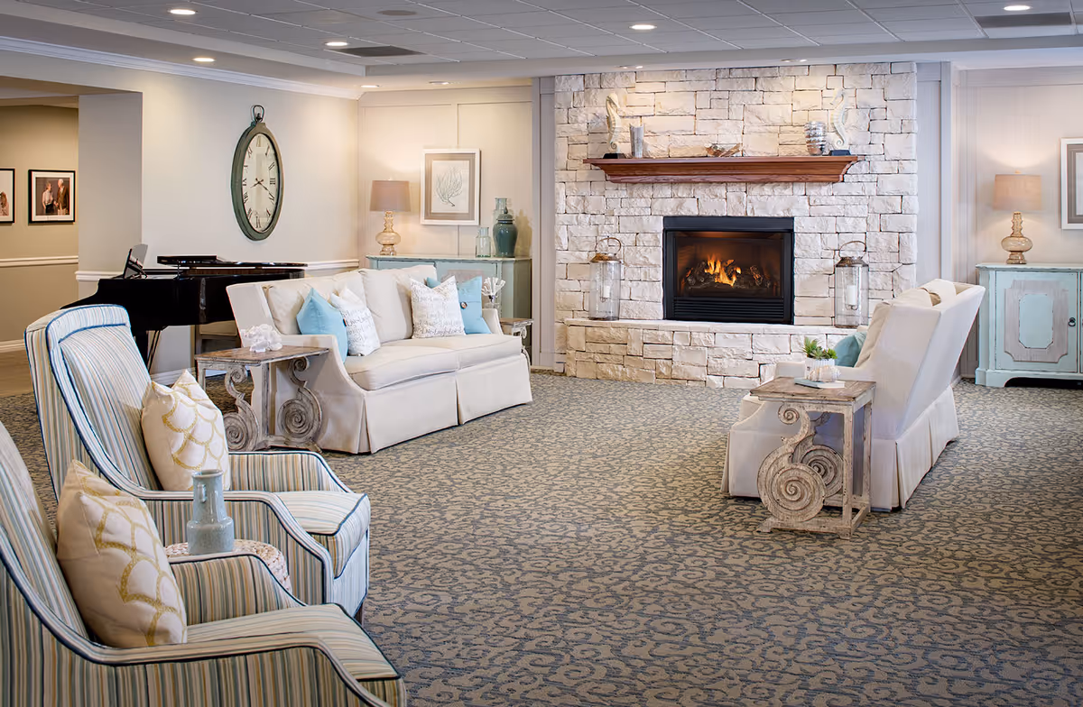 A cozy senior living room with a stone fireplace in the center, flanked by two white sofas with blue and white pillows. There are two striped armchairs with decorative pillows in the foreground. The room features a patterned carpet, a large wall clock, framed artwork, and a black grand piano in the background. Soft lighting and decorative vases add to the warm ambiance.