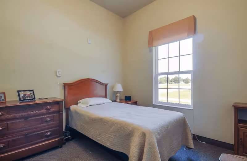 A simple bedroom in an assisted living facility featuring a single bed with a white bedspread, a wooden headboard, a wooden dresser with framed photos, a nightstand with a lamp, and a window with a peach-colored valance letting in natural light.