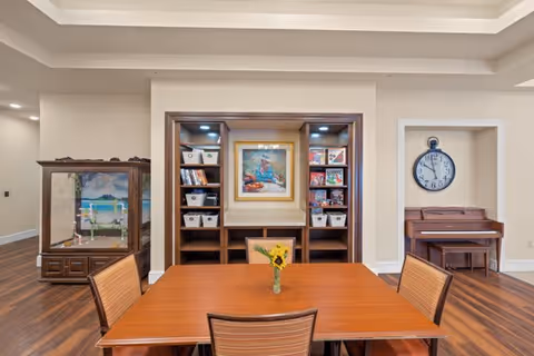 A cozy interior room featuring a wooden table with four chairs and a small vase with sunflowers in the center. Behind the table is a built-in wooden shelving unit with books, baskets, and a framed painting. To the left, there is a glass display case with a model or diorama inside. To the right, a wooden piano sits beneath a large wall clock. The room has warm wood flooring and soft beige walls.