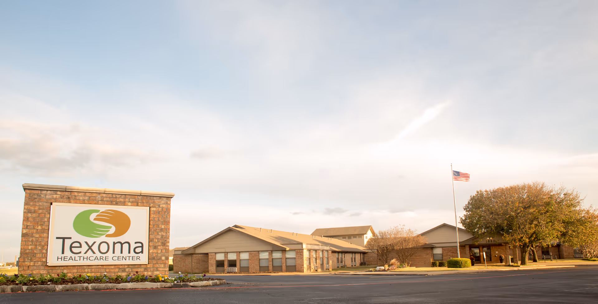 Front exterior of Texoma Healthcare Center showing a large stone sign, the low brick facility building, and an American flag under a wide sky.