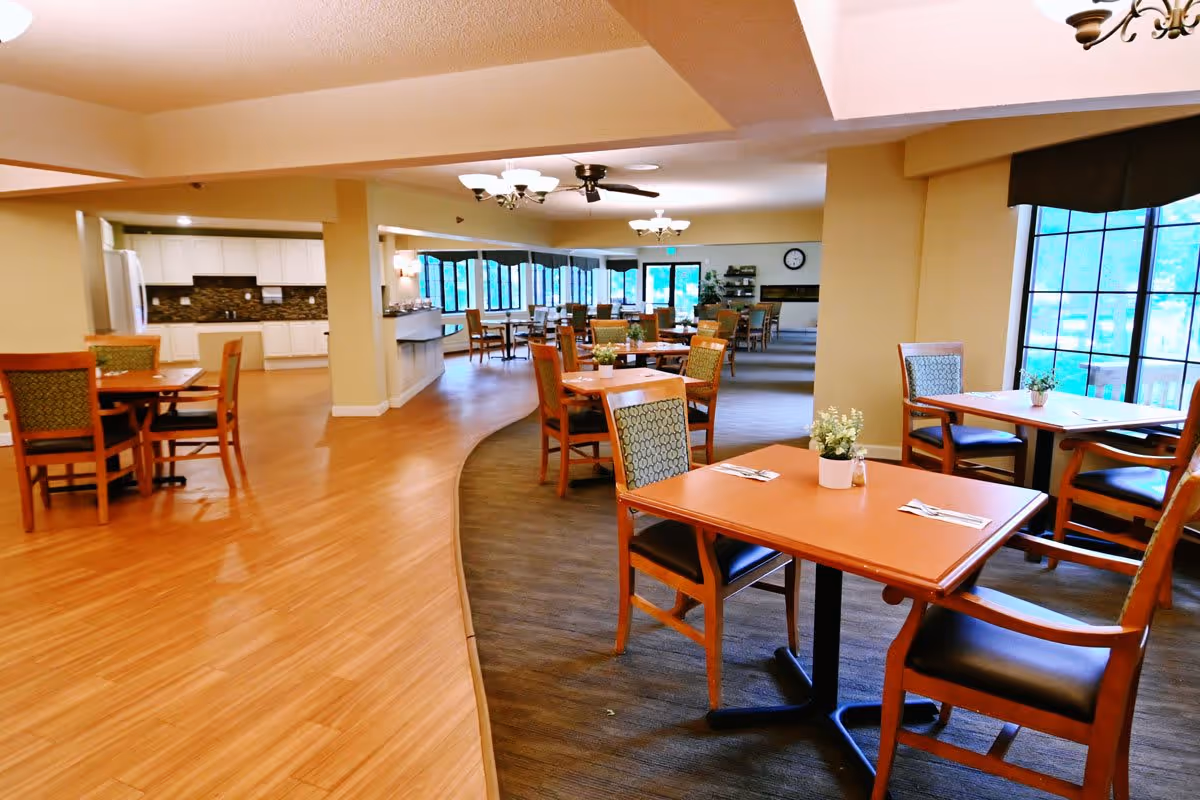 A spacious dining area in a senior living facility with multiple wooden tables and chairs arranged neatly. The room features large windows allowing natural light to brighten the space, light-colored walls, and a combination of wood and carpet flooring. There is a kitchen area visible in the background with white cabinets and a countertop.