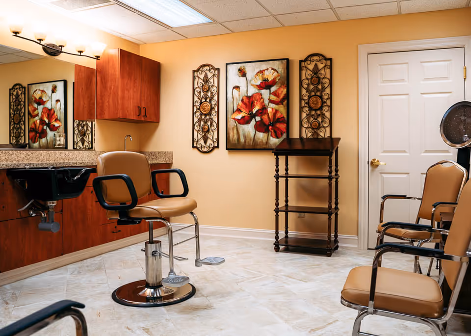 Interior of a salon room with a brown salon chair in front of a black sink and wooden cabinets. There are three additional brown chairs with metal arms, a wooden shelving unit, and decorative wall art featuring red flowers and ornamental metal designs. The room has beige walls and a tiled floor.