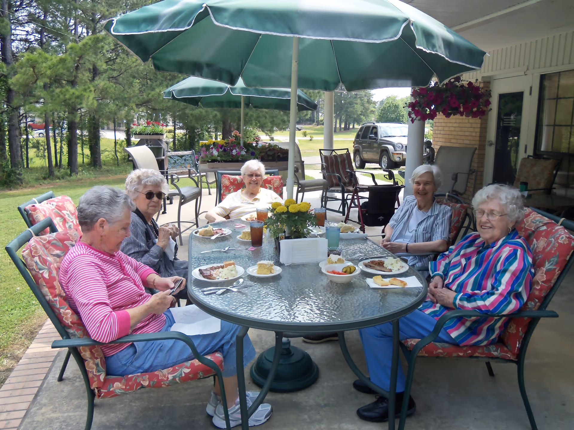 Five elderly women sitting around a glass outdoor table with floral cushioned chairs under green umbrellas, enjoying a meal together on a patio with trees and a parked car in the background.