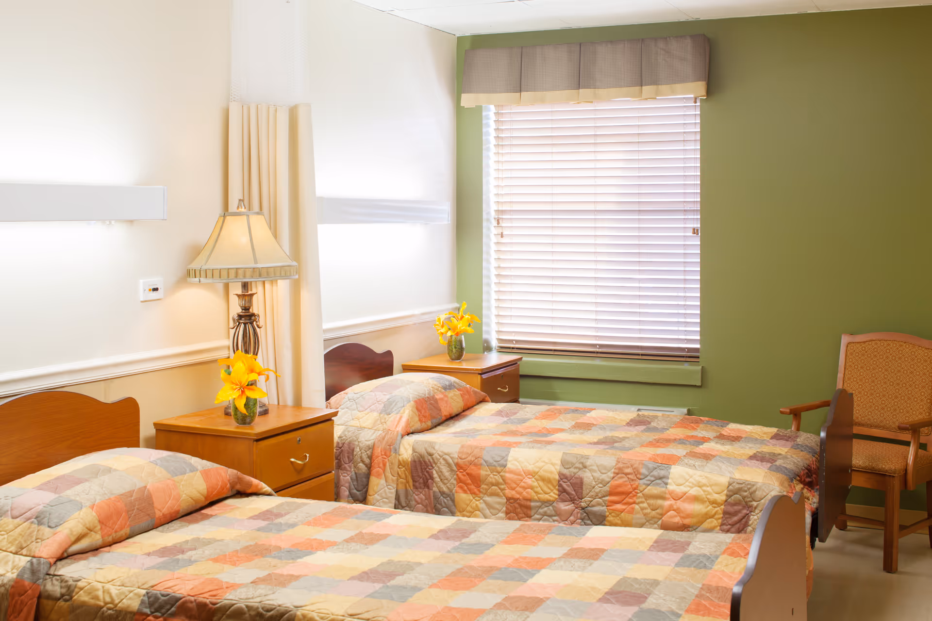 A bedroom with two single beds covered in multicolored patchwork quilts. Between the beds is a wooden nightstand with a lamp and a small vase of yellow flowers. A window with closed blinds and a beige valance is on the green wall behind one bed. An upholstered wooden chair is positioned near the window.