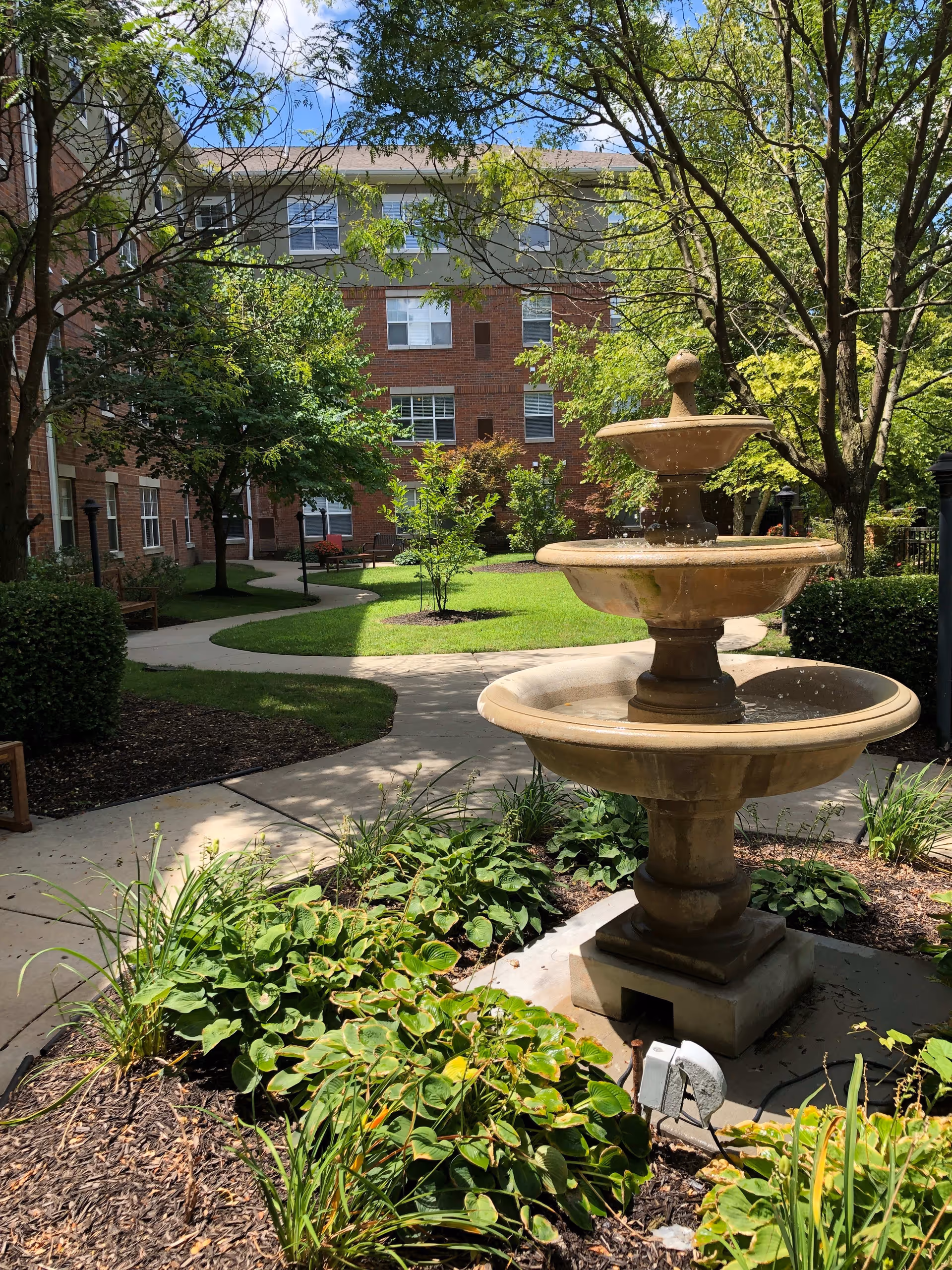 A sunny outdoor courtyard at a senior living facility featuring a three-tiered stone fountain surrounded by green plants and bushes. Curved concrete pathways wind through the landscaped garden with trees and grass, and a multi-story brick building is visible in the background under a blue sky with some clouds.
