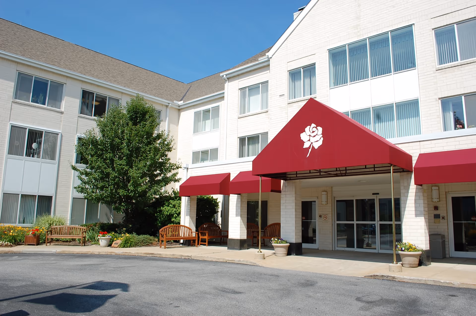 Front entrance of a light-colored multi-story senior living building with red awnings, benches, and potted plants.