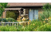 A garden area in front of a brick building featuring a multi-tiered stone fountain surrounded by various green plants and flowers, with a wooden pergola and a sliding glass door in the background.