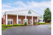Front entrance of a brick senior living facility with white columns, a covered porte-cochère, landscaped shrubs, and a paved driveway.