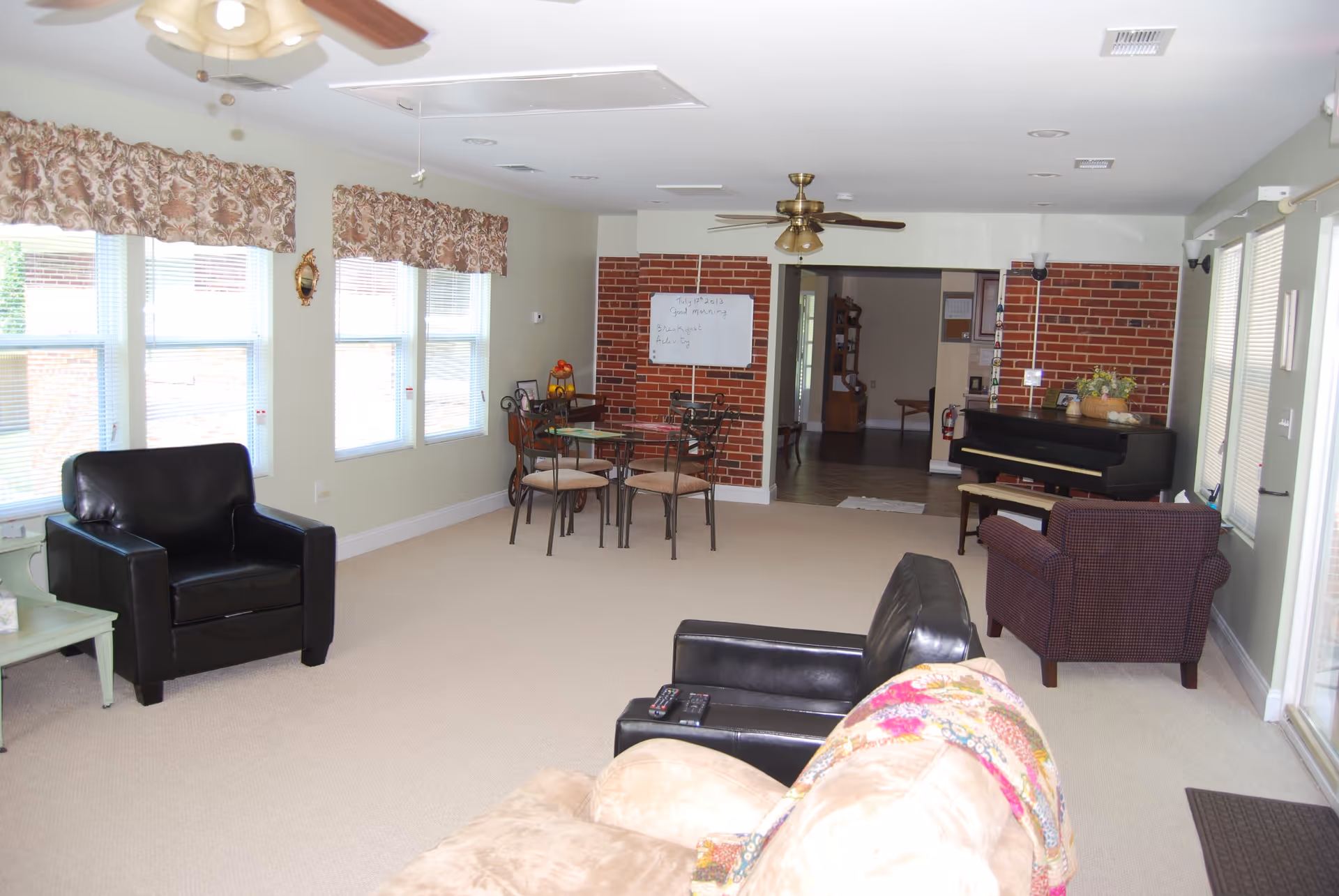 A bright living room with multiple seating options including a black leather armchair, a patterned armchair, and a beige sofa with a colorful throw. There is a glass-top dining table with four chairs near a brick accent wall. A black piano is placed against the brick wall, and large windows with floral valances allow natural light to fill the room. Ceiling fans and light fixtures are visible on the ceiling.
