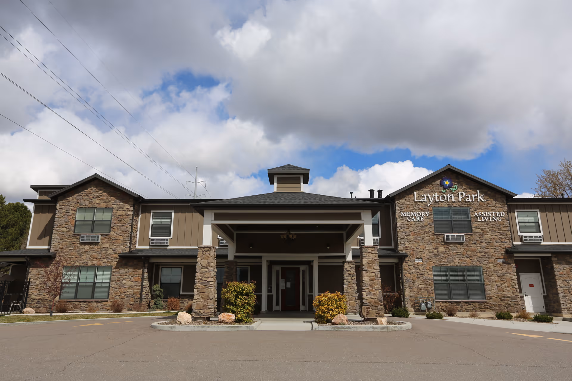 Front exterior view of Layton Park Memory Care and Assisted Living facility with stone and siding facade, a covered entrance supported by stone pillars, and a partly cloudy sky overhead.
