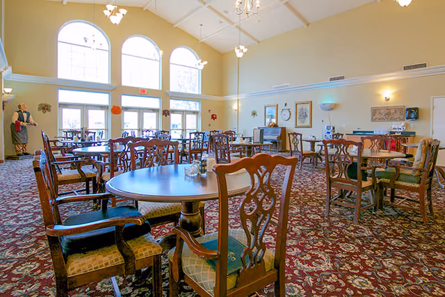 A spacious dining room with multiple wooden tables and chairs arranged on a patterned carpet. Large arched windows allow natural light to fill the room. The walls are decorated with framed artwork and a clock. A statue is visible near the entrance on the left side.