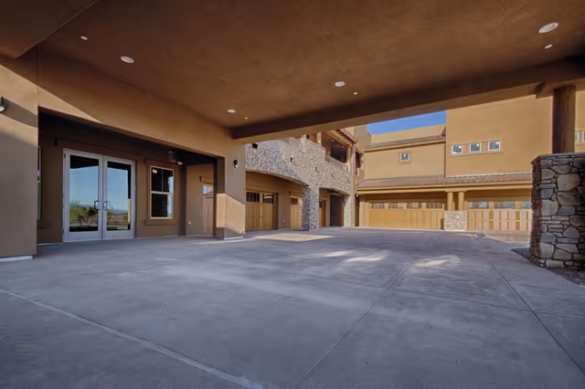 Covered outdoor area with concrete flooring and stone pillars, adjacent to a building with multiple garage doors and windows under a clear blue sky.