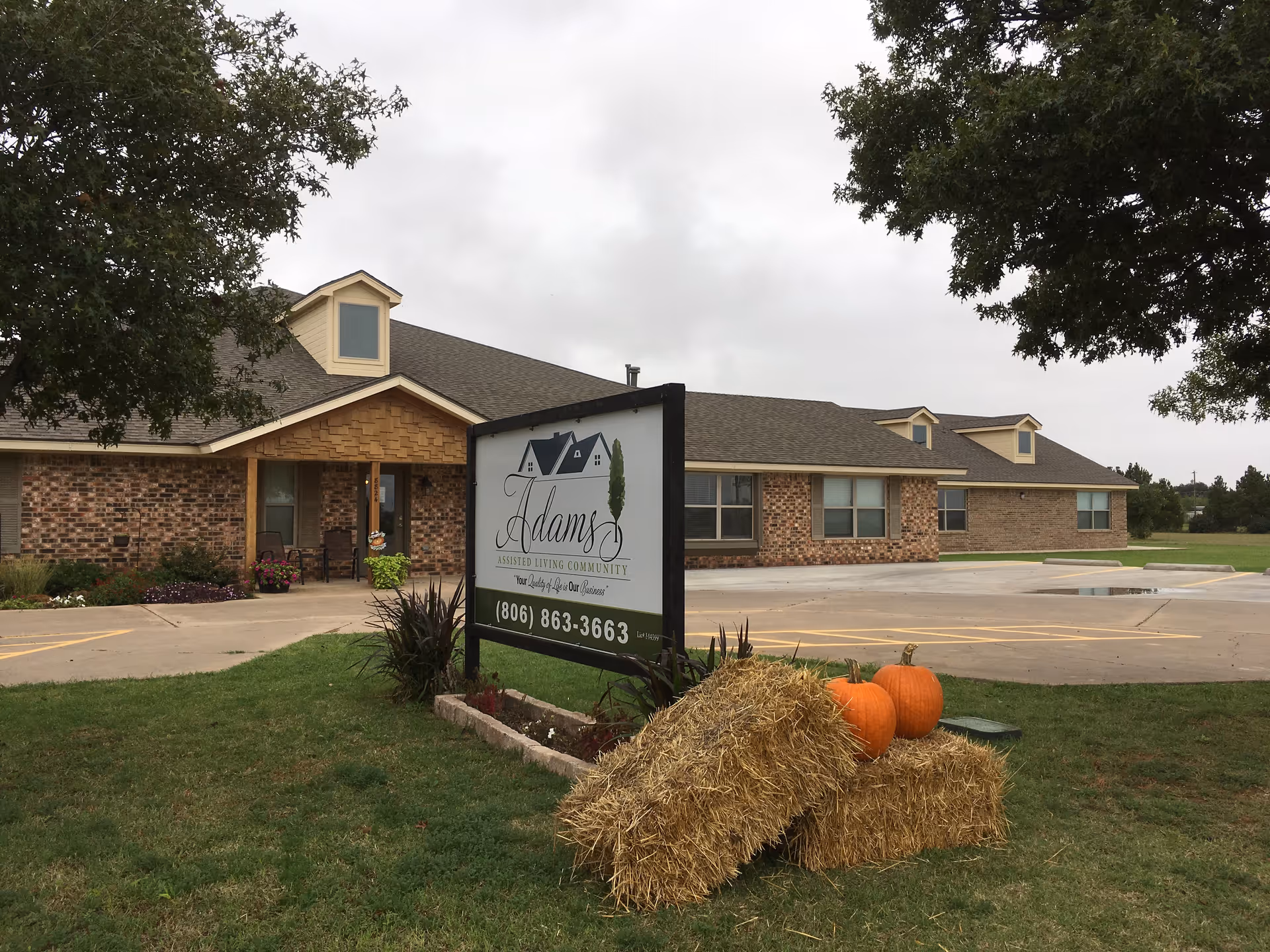 Exterior view of Adams Assisted Living Community building with a brick facade and a pitched roof. In front of the building is a sign displaying the facility's name and phone number. There are hay bales and pumpkins placed on the grass near the sign, and trees frame the scene under a cloudy sky.