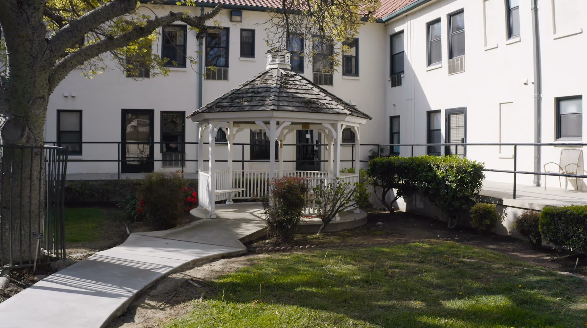 Outdoor courtyard area with a white gazebo surrounded by bushes and a tree, adjacent to a white building with multiple windows and a ramp leading to the entrance.