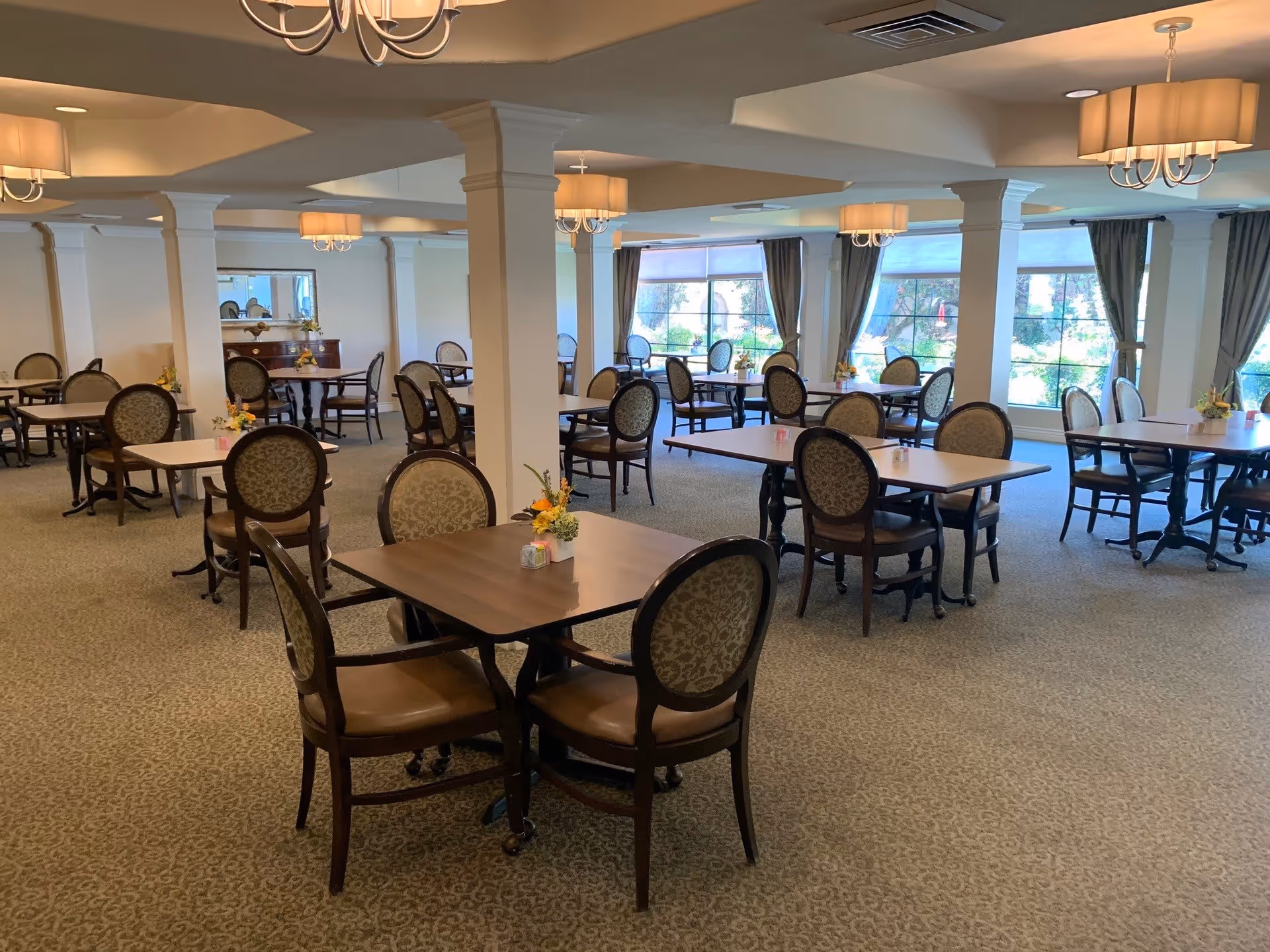A spacious dining room with multiple square wooden tables, each surrounded by four upholstered chairs with patterned backs. The room has large windows with curtains allowing natural light to brighten the space. There are several chandeliers hanging from the ceiling, and small floral centerpieces are placed on each table.