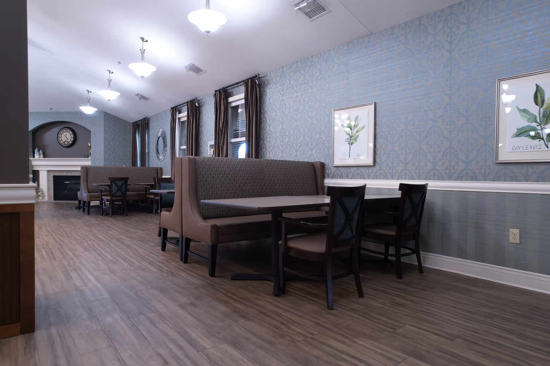 Interior view of a senior living facility dining area with booth seating and chairs around tables. The room has wood flooring, blue patterned wallpaper, framed botanical prints on the wall, and a fireplace with a clock above it in the background. Ceiling lights illuminate the space.