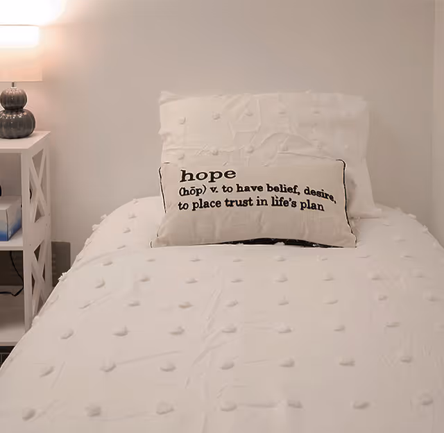 A neatly made single bed with a white textured comforter and a matching pillow. On the bed is a decorative rectangular pillow with the word 'hope' and its definition printed on it. To the left of the bed is a white nightstand with a lamp and some items on the shelves.