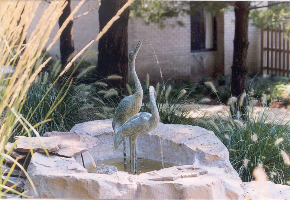 Courtyard stone fountain with two bird statues spouting water, surrounded by ornamental grasses and trees with a building in the background.